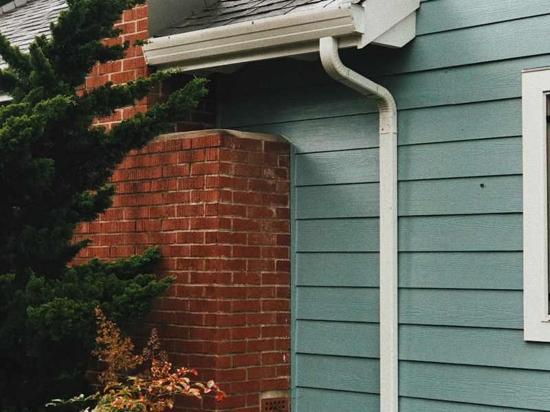 A close-up of a house corner with teal siding, white gutter, and red brick chimney.