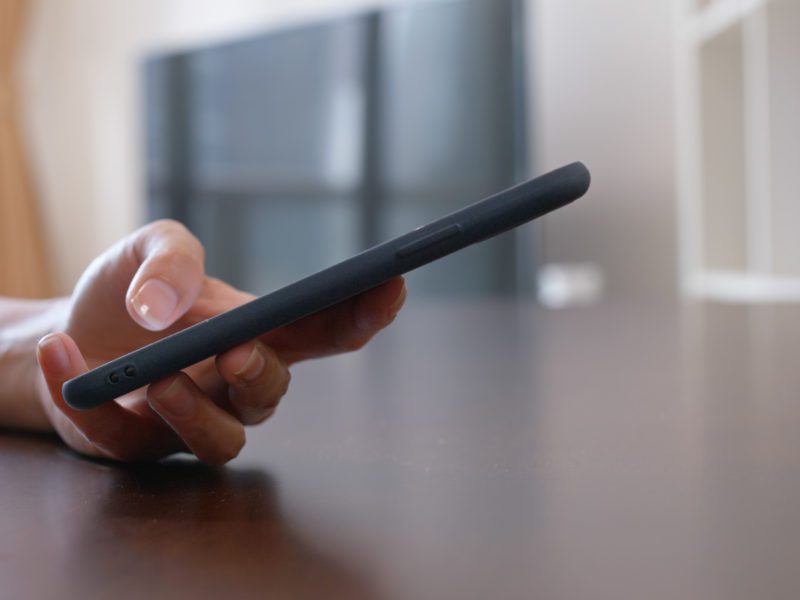 A hand grips a black smartphone resting on a wooden surface with a blurred background.