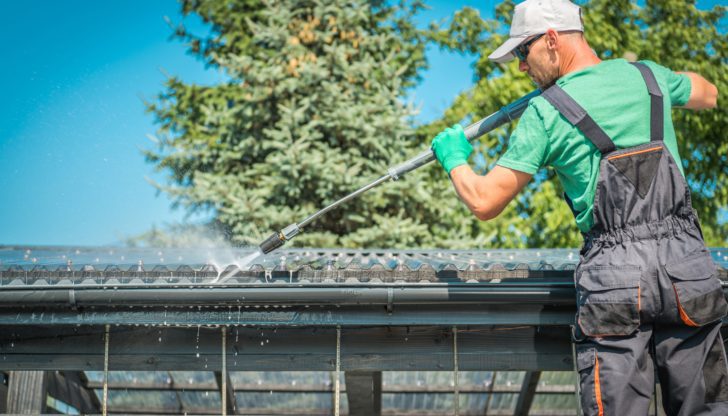 A man in work overalls uses a pressure washer to clean a roof gutter on a sunny day.
